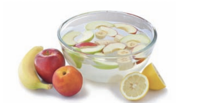 Cut apples, bananas, and lemons in a bowl of water with whole apples, bananas, and lemons next to the bowl.