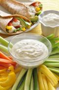 Small bowl of creamy dip with a plate of sliced vegetables and served in a veggie wrap.