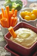 Bowl of hummus with a lemon slice served with carrot sticks, tomatoes and broccoli.