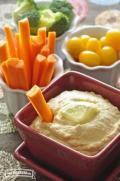 Bowl of hummus with a lemon slice served with carrot sticks, tomatoes, and broccoli.