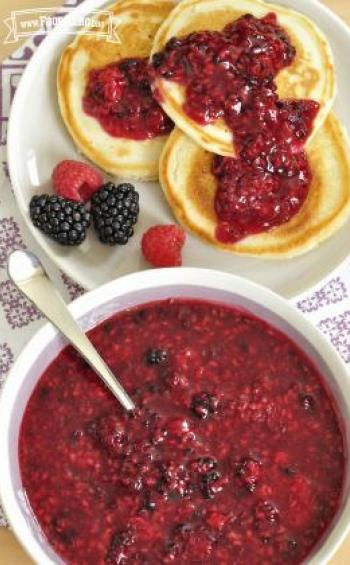 A serving bowl of Any Berry Sauce is shown with a plate of 3 pancakes that have sauce spread on top and a few fresh berries on the side.